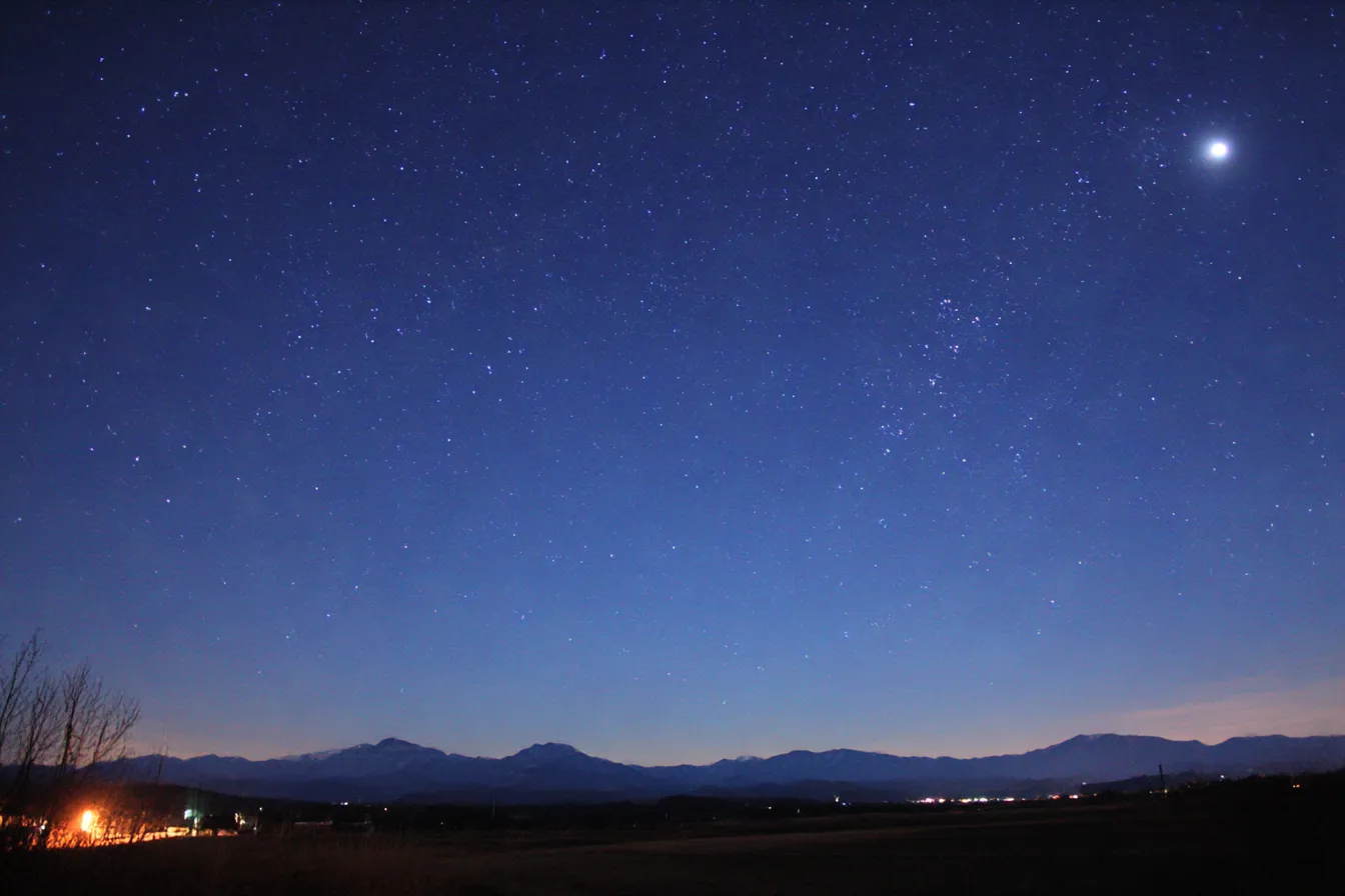 満天の星空と綺麗背景｜夜空を見上げると、無数の星が輝いています。星々と山々が静かな夜空に広がり、遠くの街灯の光がほんのりとした温かさを加え、綺麗背景がその壮大さを際立たせています。