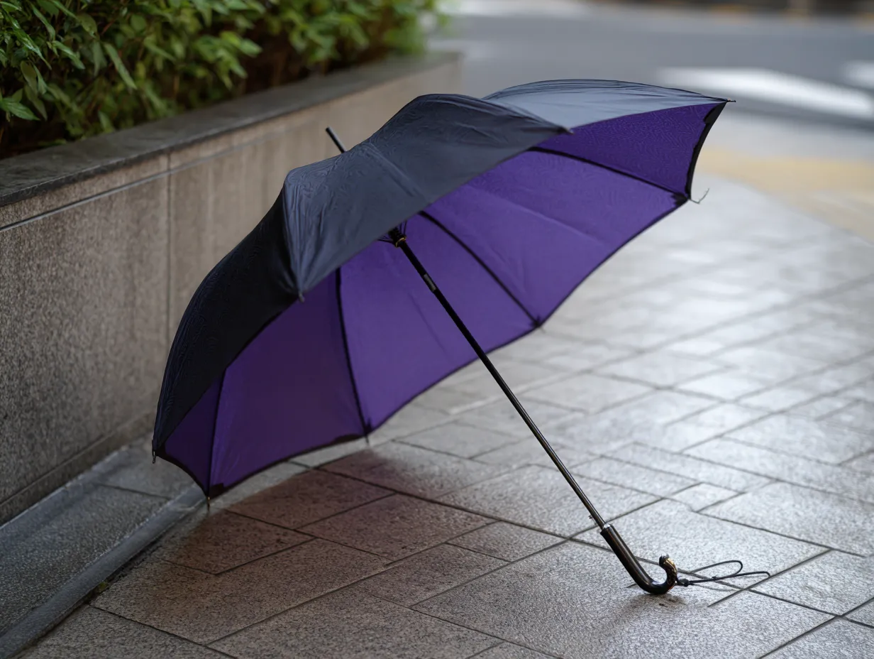 紫に合う色×傘と雨の舗道風景｜鮮やかな紫の内側と黒の外面が対照的な雨傘。濡れたアスファルトと緑の植え込みが背景に映え、しっとりとした詩的な情景を生み出している。