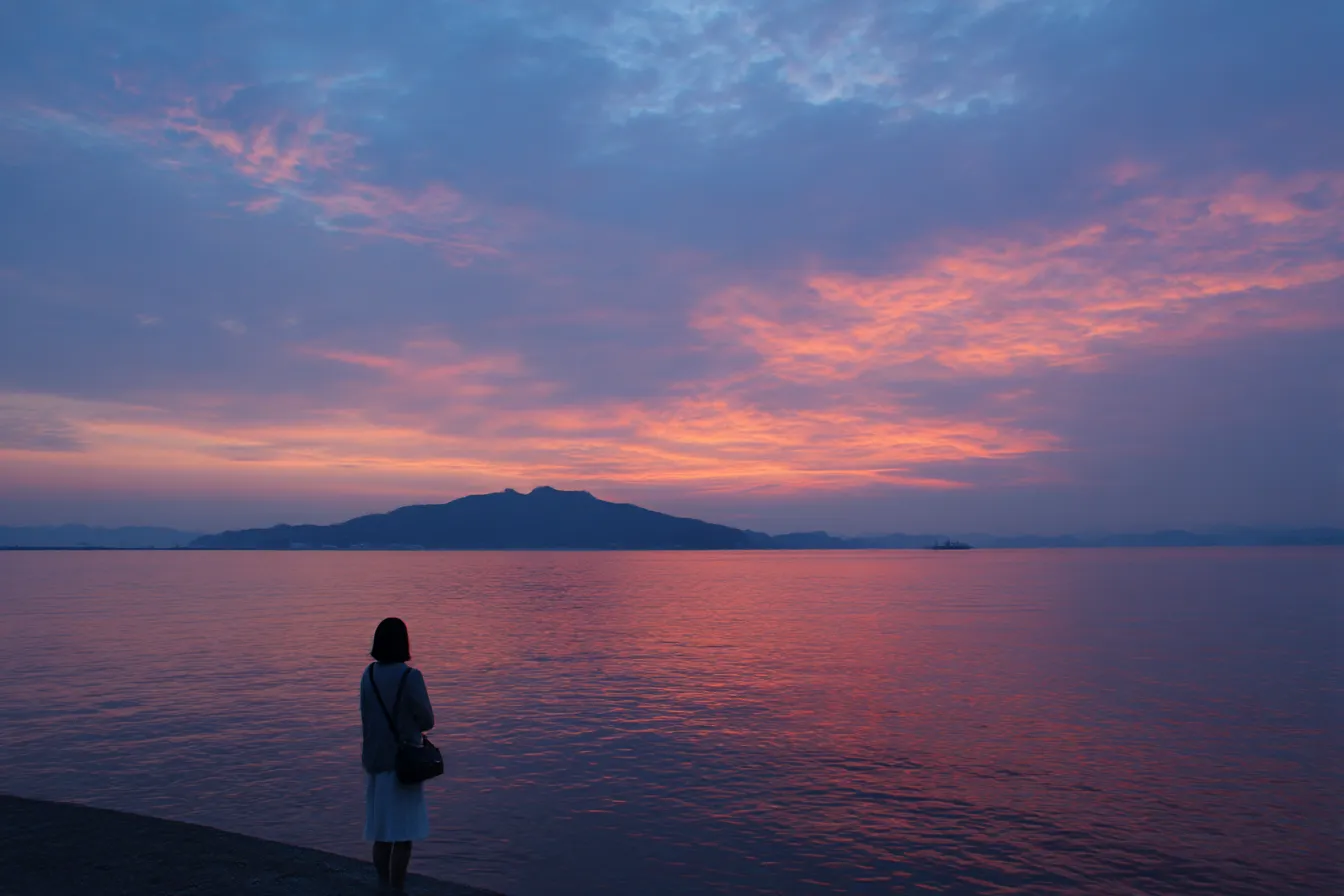 儚い背景・夕暮れの海と少女｜紫と朱が混ざる夕焼け空と海のグラデーションが美しい背景。背を向けた少女の姿が物語性を帯び、しんとした儚さを印象づける。