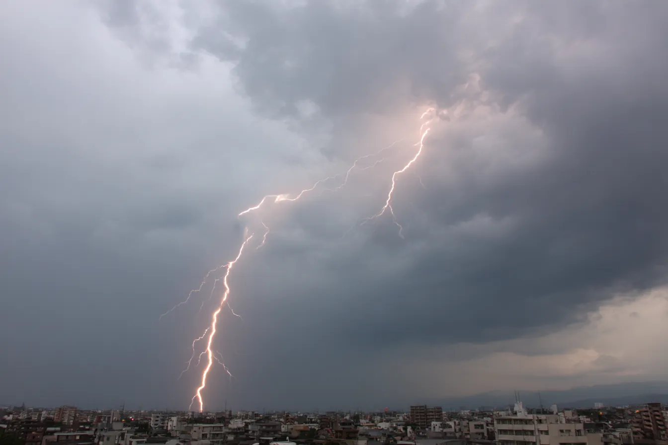 雷背景×灰空に落ちる一本の雷｜厚い雲に覆われた灰色の空から一直線に地上へ落ちる稲妻。都市のスカイラインが見えるリアルな構図で、天候描写や災害シーンにも使えるリアリティ重視の背景。