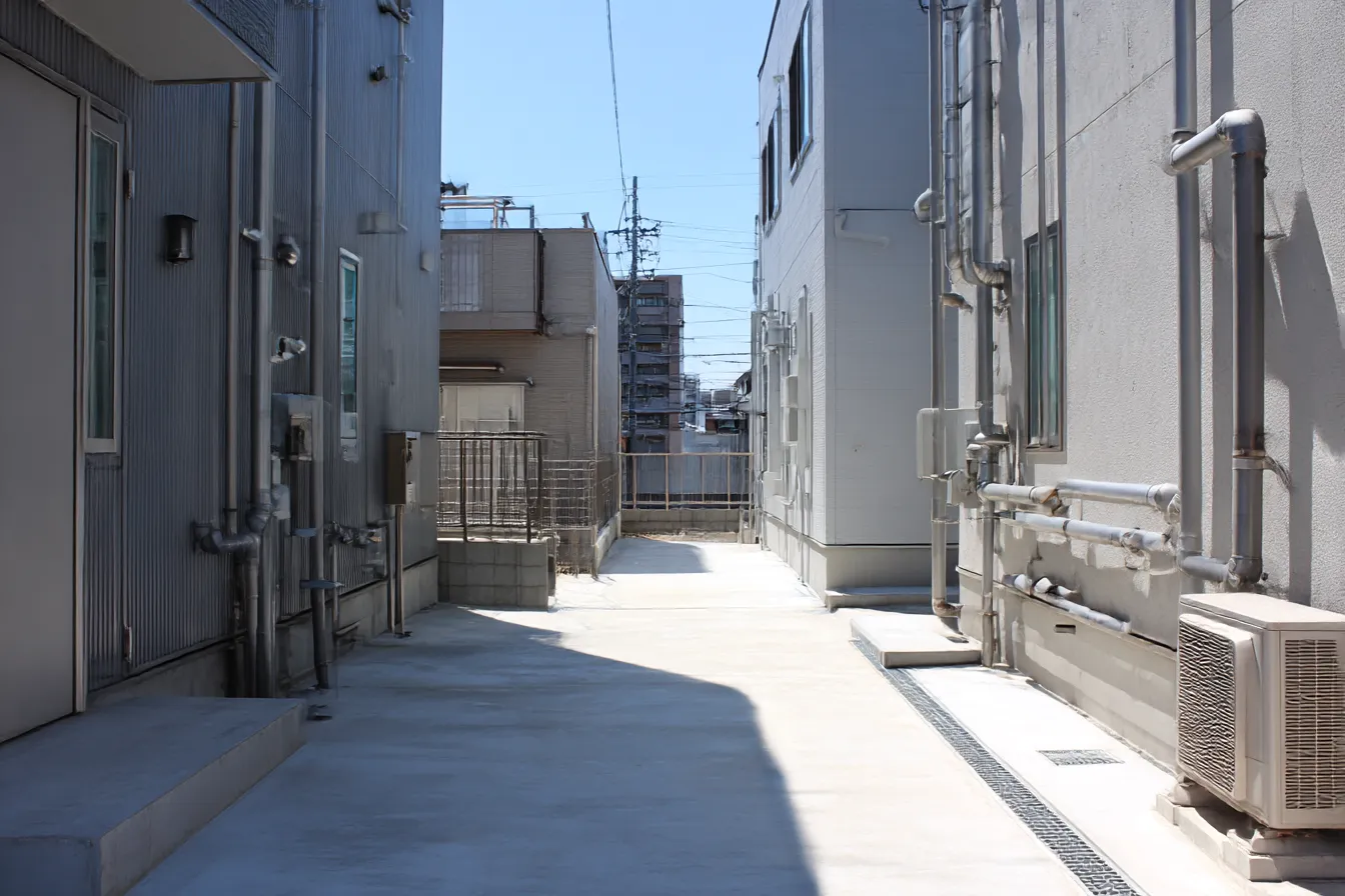路地裏背景　昼下がりの裏路地通路｜無機質な建物に囲まれた狭い裏路地が明るく照らされる背景。日差しと陰影の対比がリアルで、日常の一コマを切り取ったような静けさがある。