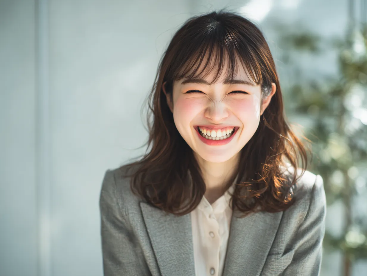 喜ぶ笑顔の女性ビジネスパーソン｜全力の笑顔を見せるスーツ姿の女性が印象的な一枚。背景の柔らかい光と明るい表情が、職場での喜ぶ気持ちを自然に引き立てます。