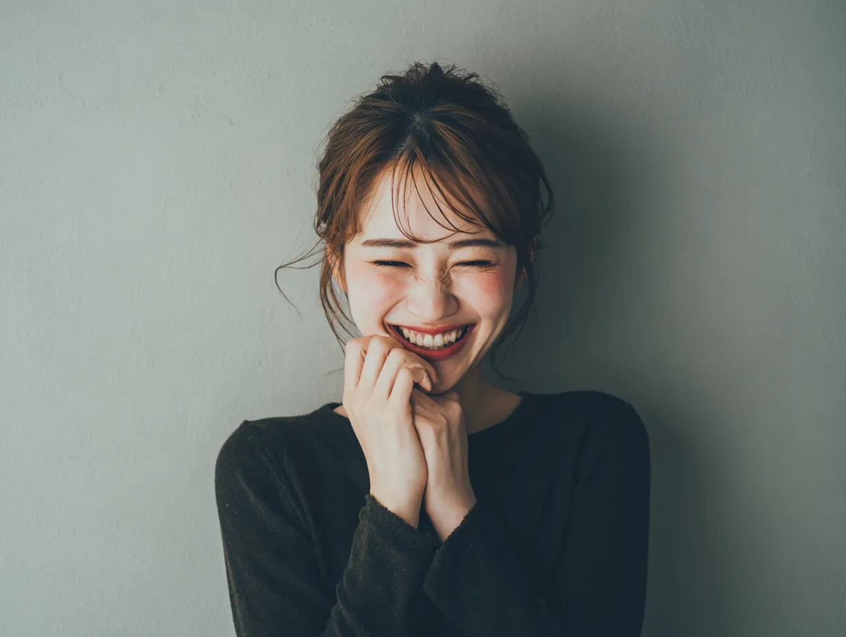 喜ぶリアクションの女性モデル｜両手をぎゅっと握ったポーズと微笑みが、嬉しさを素直に表現。背景のグレートーンが、喜ぶ気持ちとのコントラストを際立たせています。