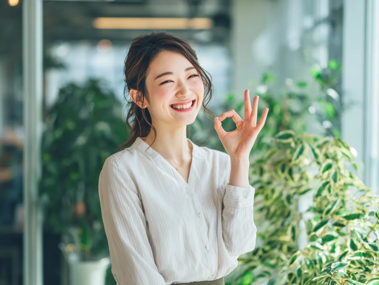 OKで安心感を伝える女性社員｜観葉植物に囲まれた柔らかな光の中で、ナチュラルな笑顔が印象的なOKサイン。信頼感やリラックスしたビジネス環境を表現する素材として効果的です。