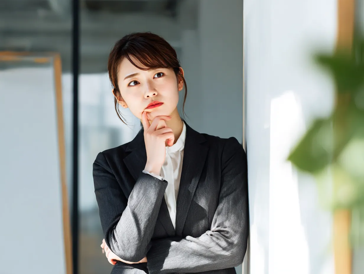 はてなを浮かべる女性社員｜自然光が柔らかく差し込むオフィスで、スーツ姿の女性が思案顔を浮かべる構図。考え込む仕草が印象的で、ビジネスシーンにおけるはてなの感情を的確に表現した素材です。