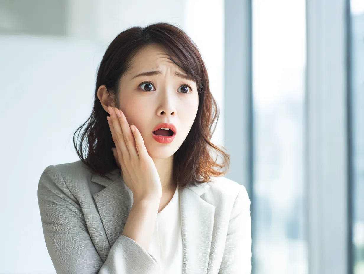 驚愕する女性ビジネスパーソン｜頬に手を当てたまま目を大きく開けて反応する女性が主役。白を基調とした清潔感ある背景が、思いがけない事態への反射的な驚きをリアルに引き立てています。
