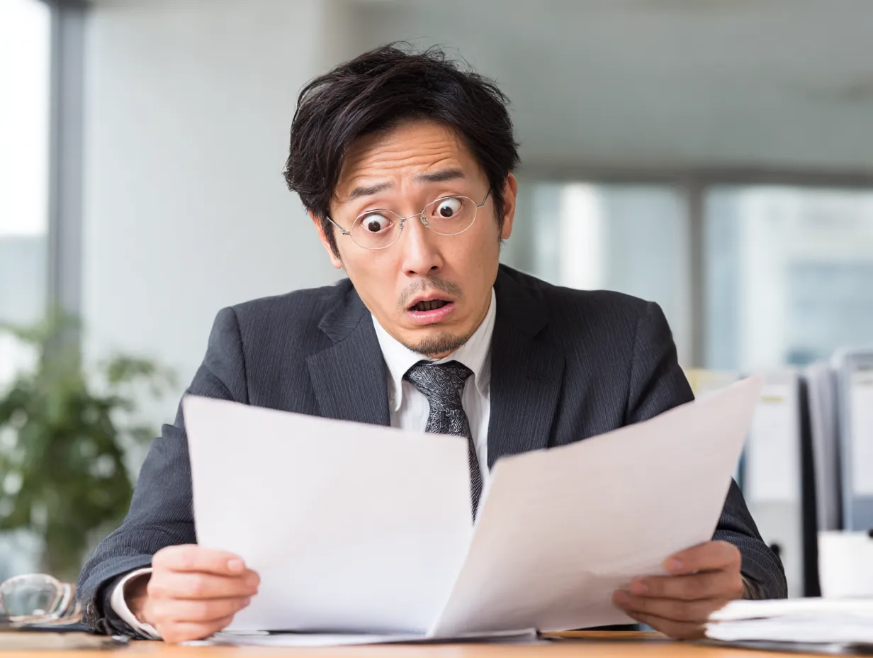 びっくりするスーツ男性の資料確認｜焦りや戸惑いのニュアンスを含んだびっくりの表情を捉えたビジネス向け素材。オフィス背景と自然光によりリアルな日常感が強く、企画やトラブルを伝えるシーンに適しています。