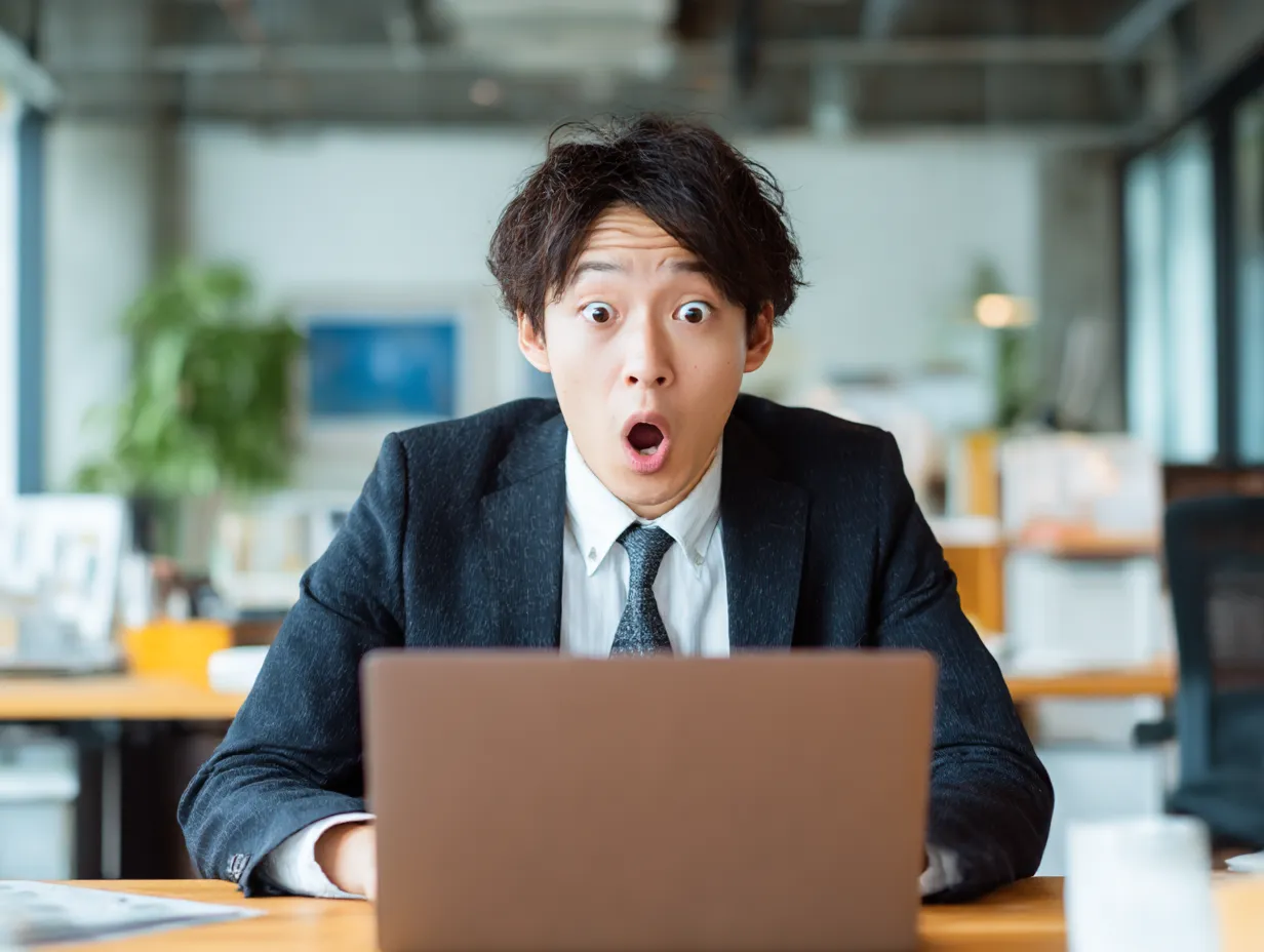びっくりした若手社員の表情（PC）｜パソコンを見て目を見開いた驚きの表情が印象的な若手社員の写真です。びっくりのシーンを自然に再現できる、ポジティブなサプライズ演出にも活用できる素材です。