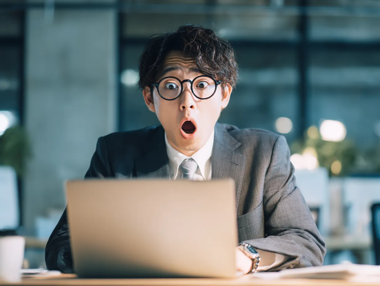 びっくり顔のメガネビジネスマン｜大きく見開いた目と口元が特徴的なびっくり顔。メガネの反射やライティングの強弱が印象的で、驚きの中に少しコミカルさも感じさせる表情が魅力のビジネス写真です。