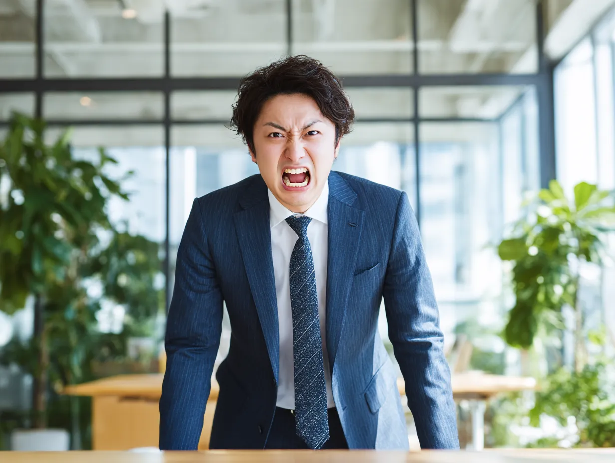 怒っているスーツ男性のビジネス表情｜机に手をついて大声で怒鳴るような表情が印象的なビジネス男性の写真です。怒りの中にびっくりも含んだ緊迫した雰囲気が伝わる構図で、トラブルシーンの表現に最適です。