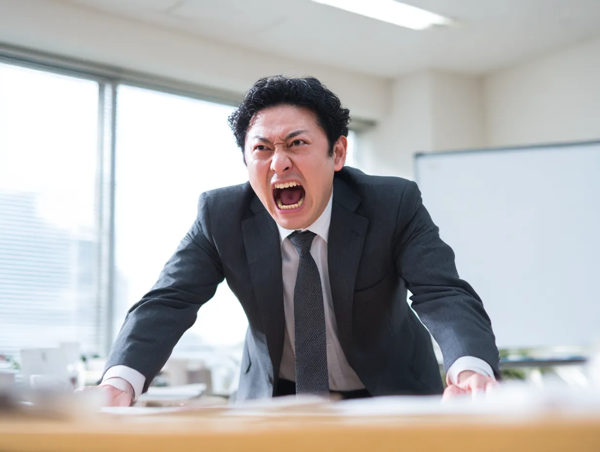 怒鳴るビジネスマン（強い感情）｜机を押さえた姿勢と全力で怒る口元が印象的な写真です。スーツ姿と整った背景がビジネスシーンの信憑性を高め、びっくりからの激昂を表現したい状況に適しています。