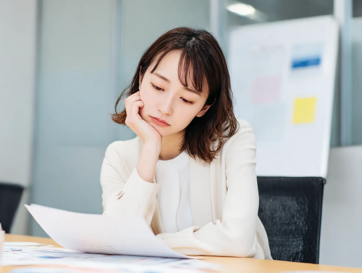 びっくりして困る女性社員の表情｜ホワイト系のスーツで書類を見つめながら頬杖をつく姿が印象的な女性の素材。びっくりした後の混乱や静かな困惑を自然な表情で表現しており、シーンの汎用性も高いです。