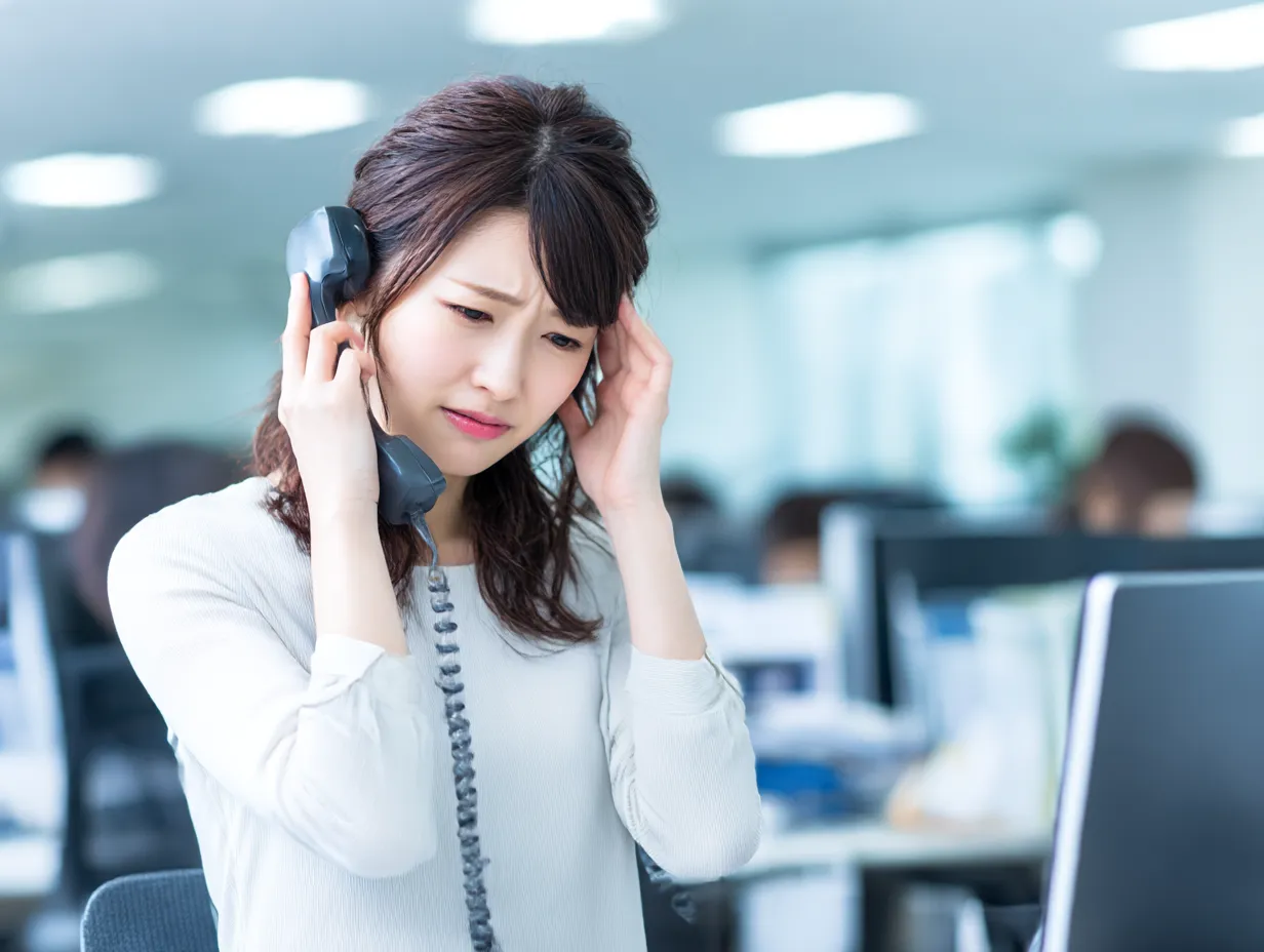 ストレスを受けた電話応対中の女性｜電話対応中の戸惑う女性がストレスを感じる様子を自然に演出。オフィスの背景と表情のギャップがリアリティを高め、顧客対応やトラブル場面を想定した素材として最適です。