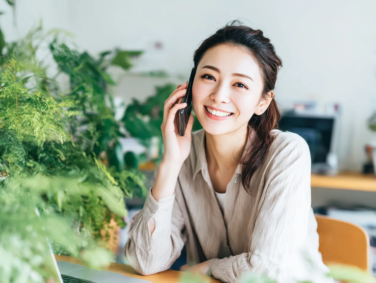 明るく電話対応する女性の笑顔｜観葉植物に囲まれた空間でスマホ通話中の女性が満面の笑みを見せる。自然光とグリーンが調和した構図が、リラックス感と信頼感を伝える明快なビジネスイメージに仕上がっています。