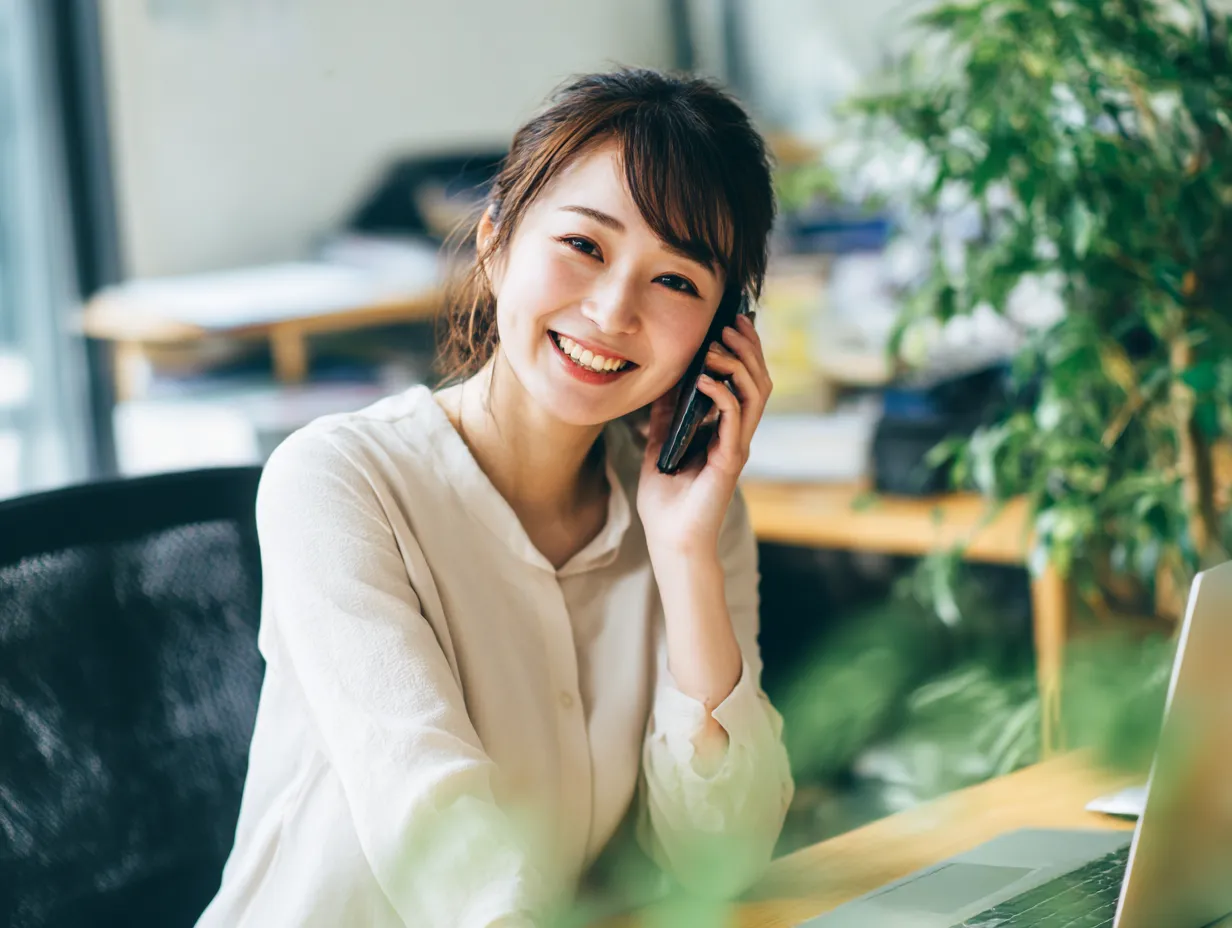 オフィスで笑顔の電話対応女性｜植物に囲まれたナチュラルな職場環境で電話を受ける女性の笑顔が印象的。親しみやすさと信頼性を感じさせる柔らかな構図で、テレワークや窓口業務の素材にも適しています。