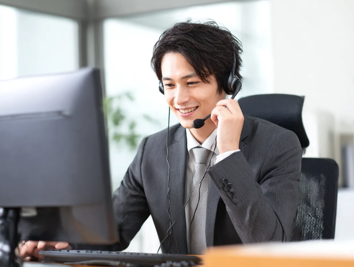 ヘッドセットで電話応対する若手男性｜オフィスでモニターを見ながら対応する男性のスマートな表情が印象的。ヘッドセット姿と整ったスーツスタイルが、コールセンターやサポート業務の信頼感を高めます。