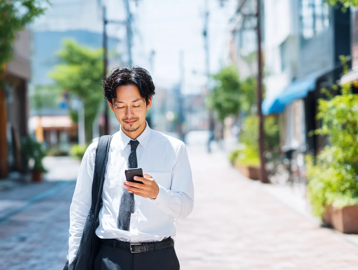 スマートフォンを使うカジュアルスーツ男性｜シャツとネクタイ姿の若手男性が、日差しの中でスマートフォンを操作。ナチュラルな構図と柔らかい表情が、親しみやすい印象をもたらす。