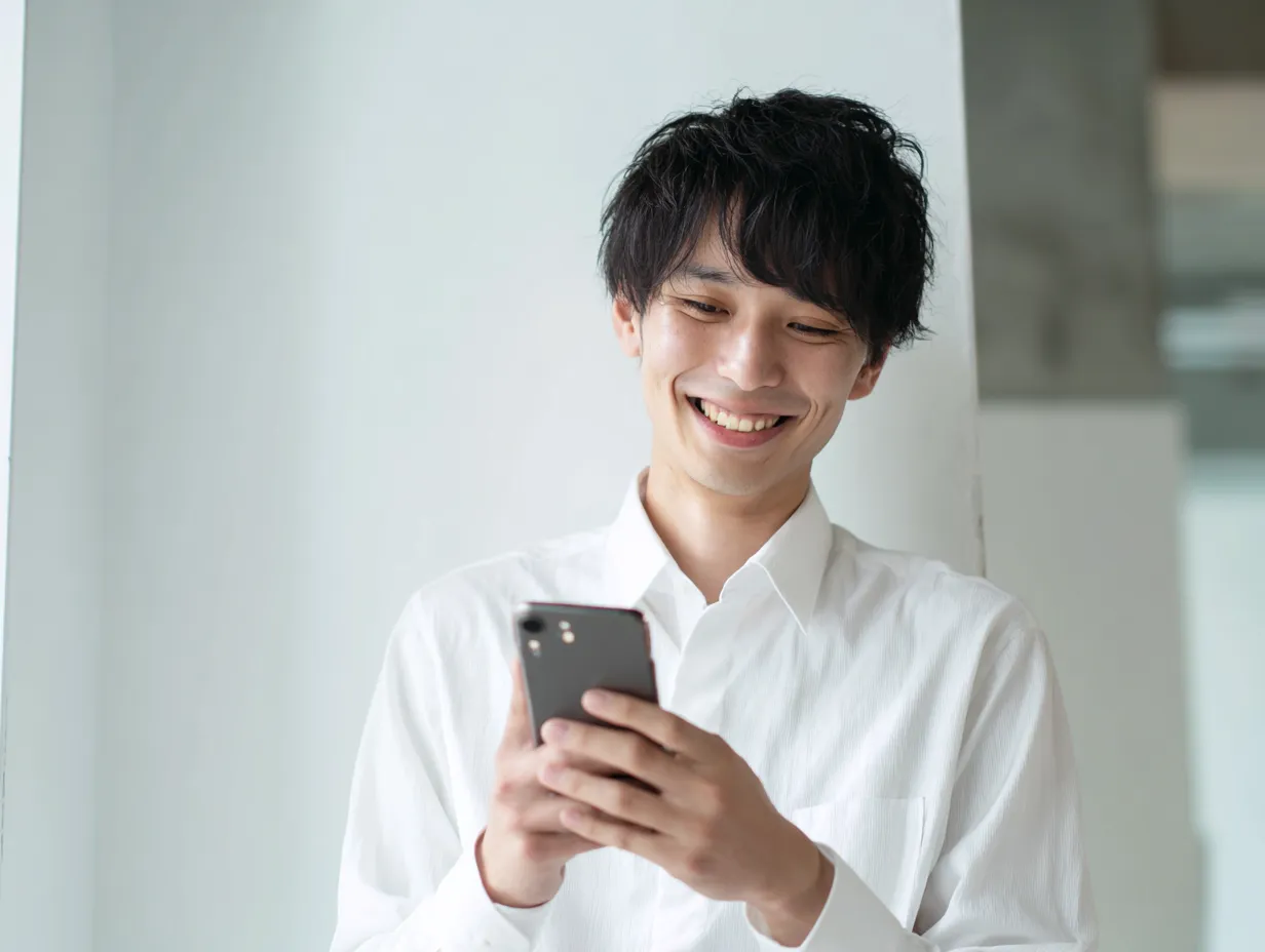 スマホを見る白シャツの若手男性｜白壁の前でスマホを見ながら微笑む若手男性。明るくミニマルな構図が、ポジティブなデジタル活用の雰囲気を引き出す現代的な一枚。