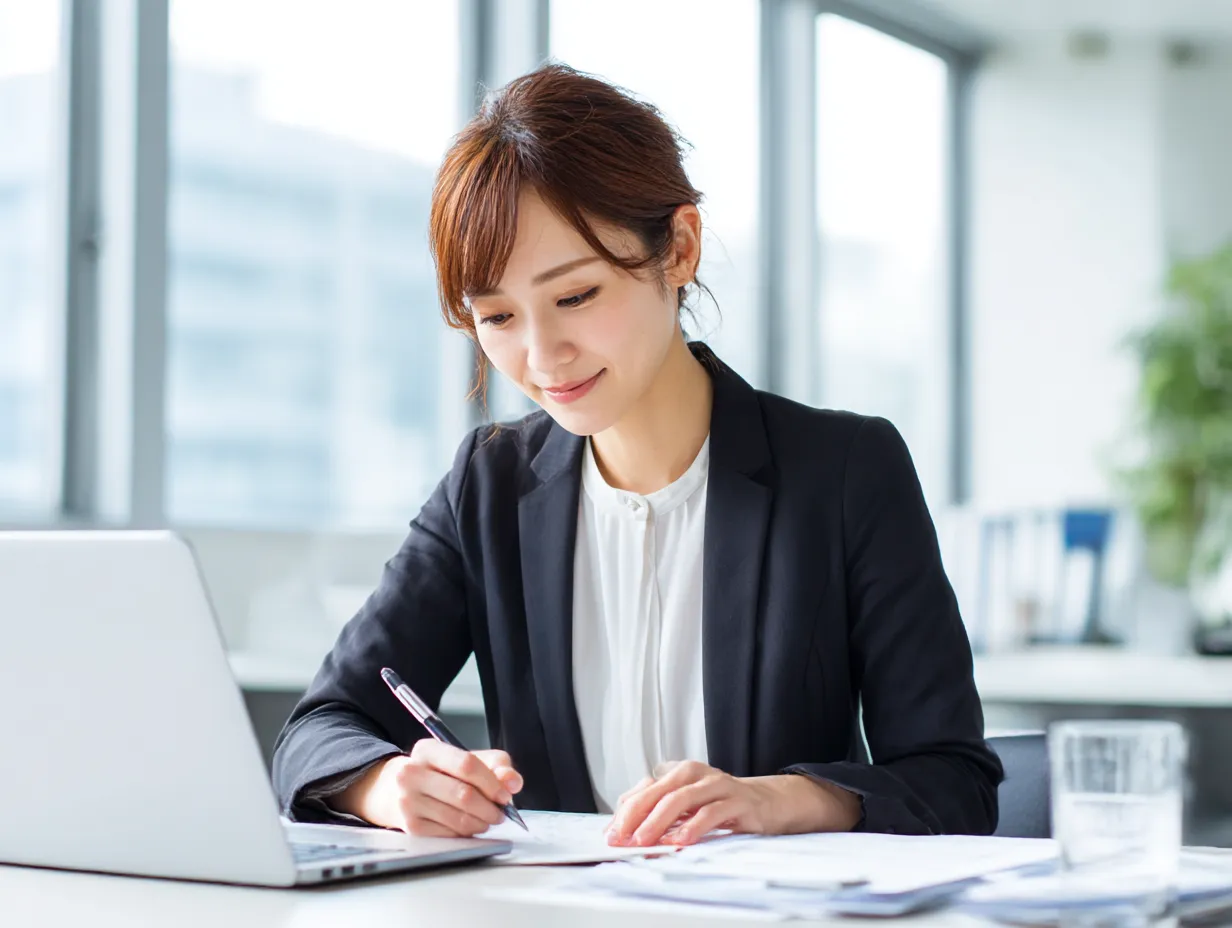 仕事中の笑顔が魅力の女性｜オフィスで資料作成をする女性の明るい表情が際立つ写真。清潔感のある配色と柔らかな光が、ポジティブで前向きな「仕事」シーンを表現している。