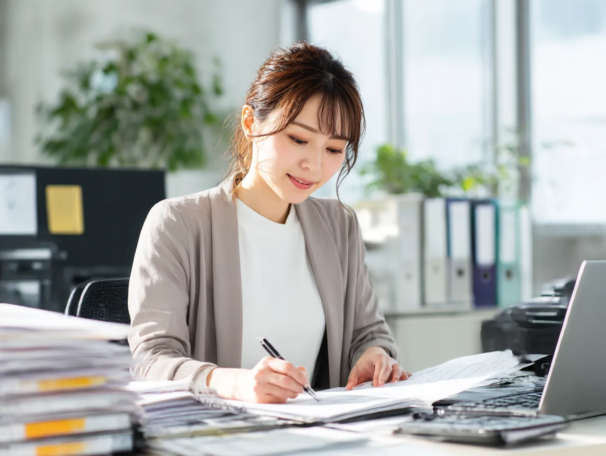 仕事に集中する女性の横顔｜日差しの差し込む明るいオフィスで書類をまとめる女性。自然な笑顔と清楚な印象で、日常の「仕事」風景をさわやかに伝えるビジネス素材。