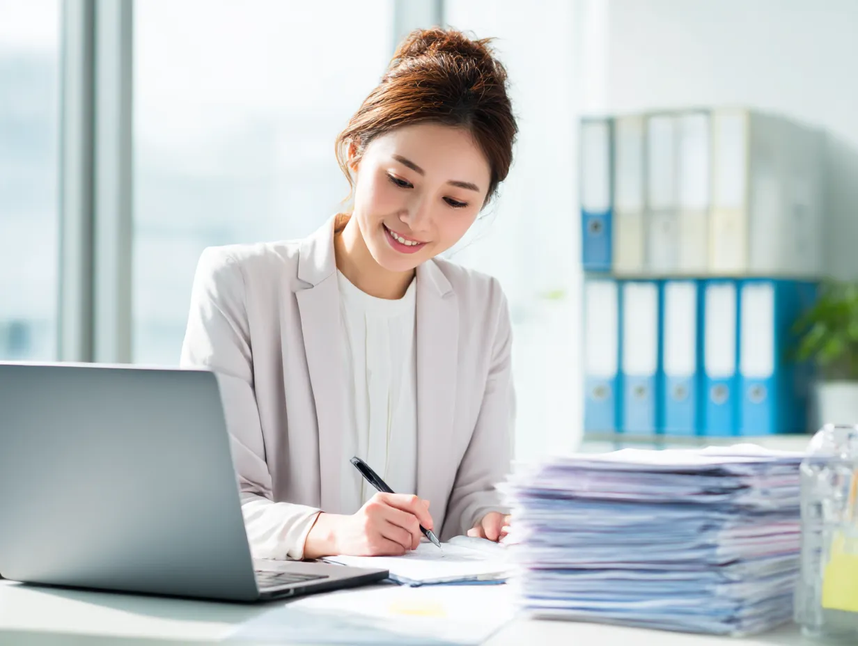仕事に取り組む女性の笑顔｜明るいオフィスで大量の書類を前に、笑顔で作業を進める女性の姿が印象的。整理された背景と清潔感あるスタイルが、前向きな仕事への姿勢を伝えるビジネス素材。