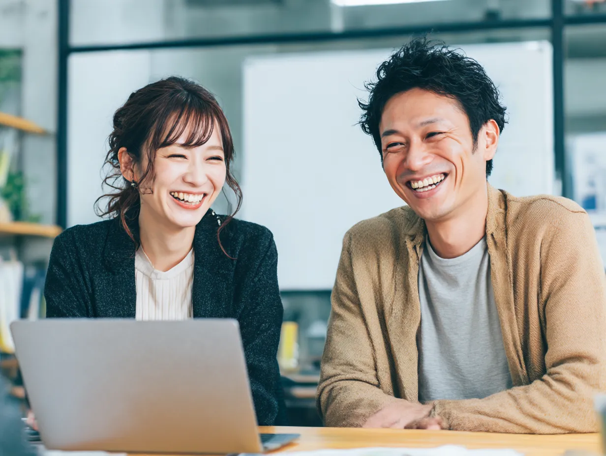 パソコンと楽しい仕事時間｜オフィスのカジュアルなミーティングシーンで、パソコンを使いながら笑顔を交わす男女。仕事を楽しんでいる雰囲気が伝わり、チームワークやコラボレーションのイメージに最適です。