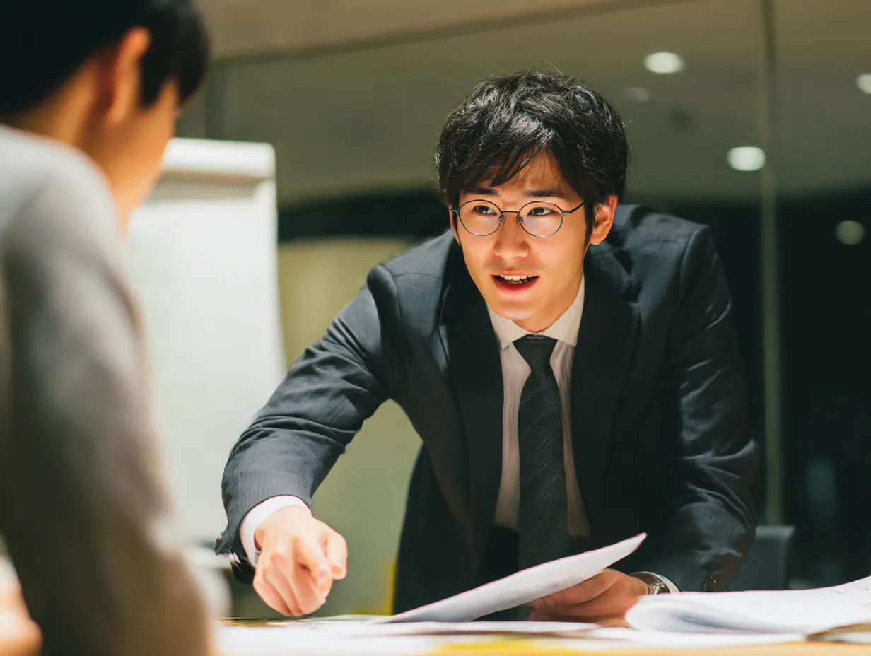 仕事の説明をしながら会話するビジネスシーン｜テーブルを囲んで資料を持ちながら、意見交換をしているシーンです。笑顔で説明しながら意見を交わす姿が、協力的で円滑なコミュニケーションを表現しています。仕事の打ち合わせや相談シーンに最適です。