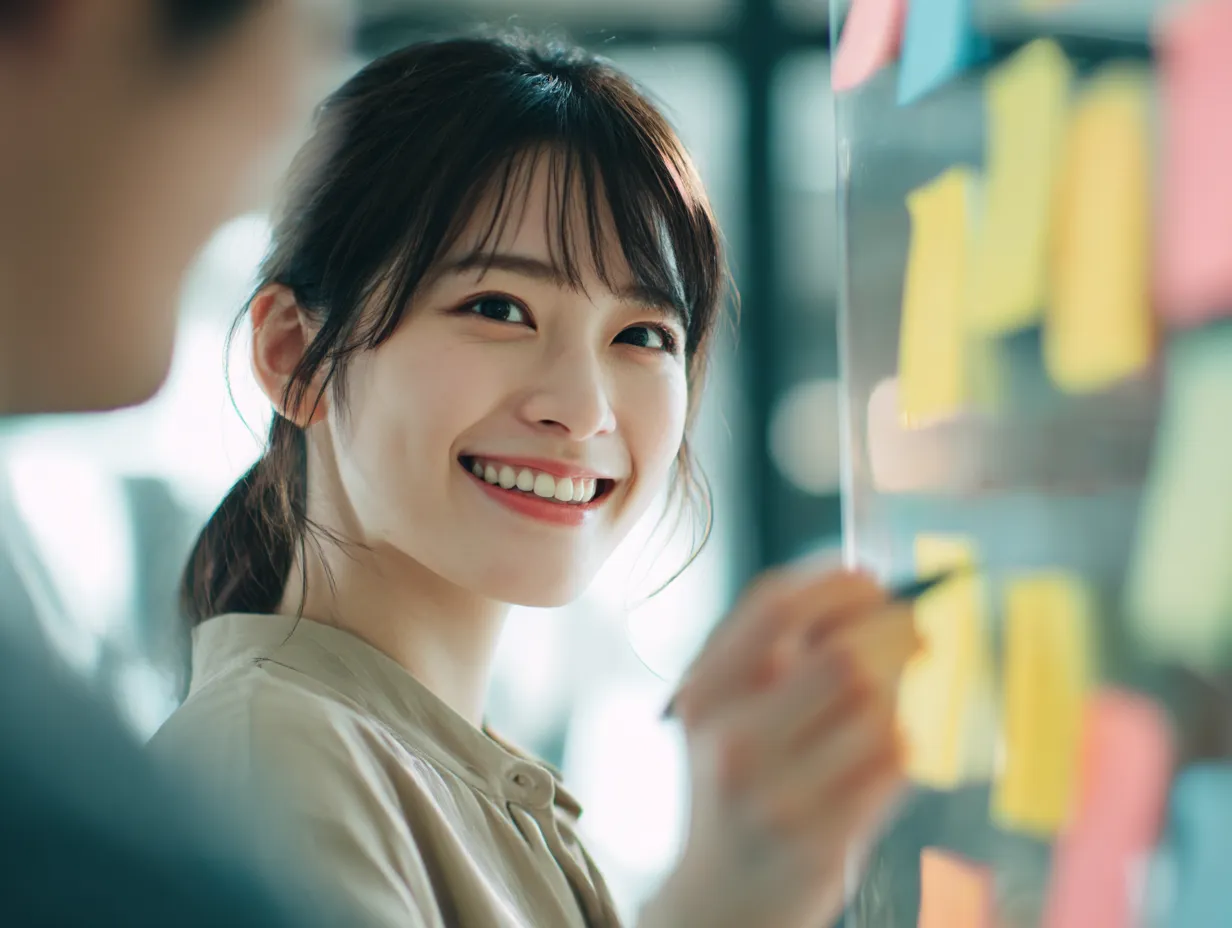 笑顔で付箋を使う女性の説明｜明るいオフィスで笑顔の女性が付箋にメモを取る様子を描いたビジネス写真素材。チーム内の「説明」を丁寧に行う場面を想起させ、親しみやすく前向きな印象を与える。