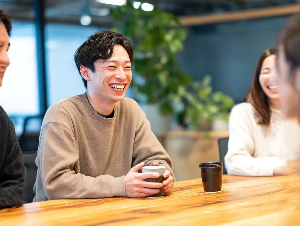 コーヒーブレイクで笑顔に話す社員の話す｜明るいオフィスでカップを片手に雑談する社員たちのシーン。リラックスした「話す」空気感がチームのつながりや社内コミュニケーションを想起させる。
