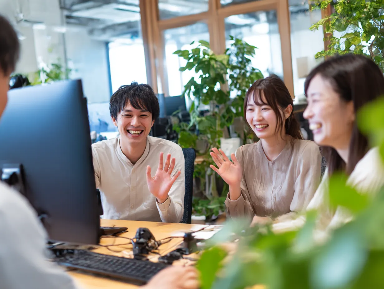 オフィスでWeb会議に参加する若手社員｜自然光と観葉植物が印象的なオフィス空間で、和やかに画面越しの相手に手を振る若手社員たちが登場し、親しみのあるWeb会議シーンを演出している。