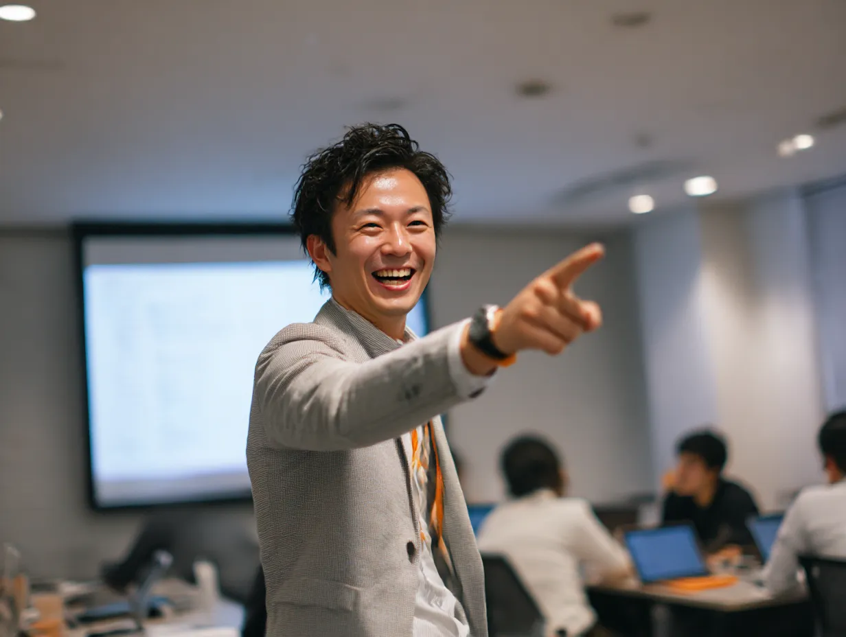 研修を盛り上げる若手講師｜エネルギッシュでフレンドリーな若手講師が指を指しながら笑顔を見せるシーン。「研修」の場面で場を和ませる指導者像を表現しており、親しみと信頼感が演出されている。