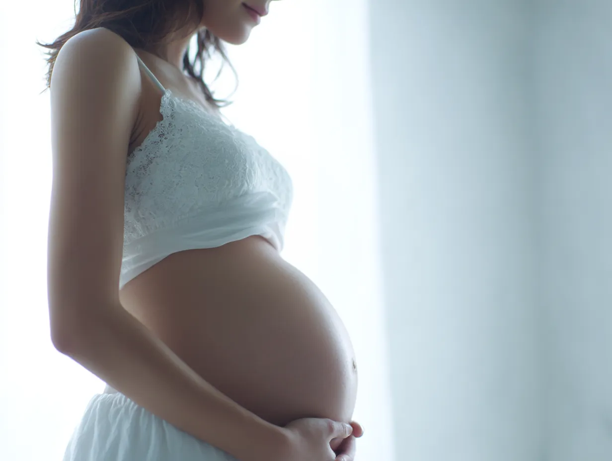 妊娠五ヶ月のおなかの輝き｜レース素材のトップに包まれた女性が光の中で妊娠五ヶ月のおなかを抱く。透ける布地と柔らかな丸みが印象的で、生命感に満ちた表情が画面に宿る。