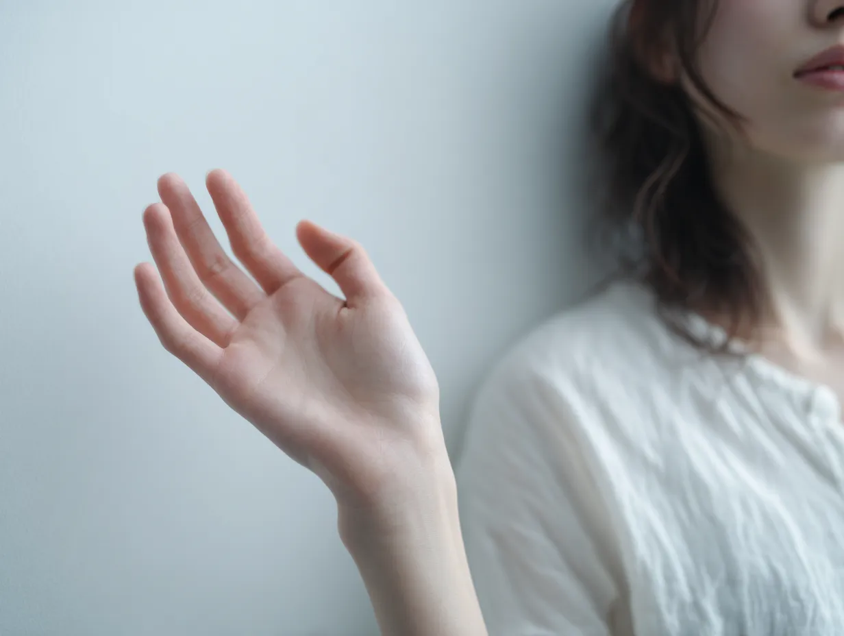 壁にかざす女性の手｜壁際に差し出された女性の手が、光を受けて浮遊感を纏う。柔らかな指のラインと口元の一部が画面に余韻を与える構成が特徴的。