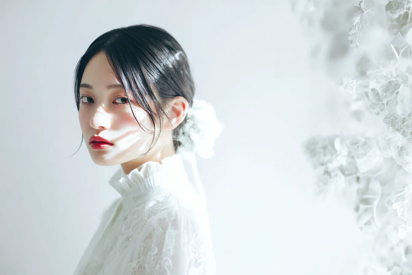 白ドレスの女性と光差す背景｜繊細なレースの白ドレスが柔らかい光に包まれ、花のような装飾が背景に溶け込む。白の静けさが空気感を引き締める。