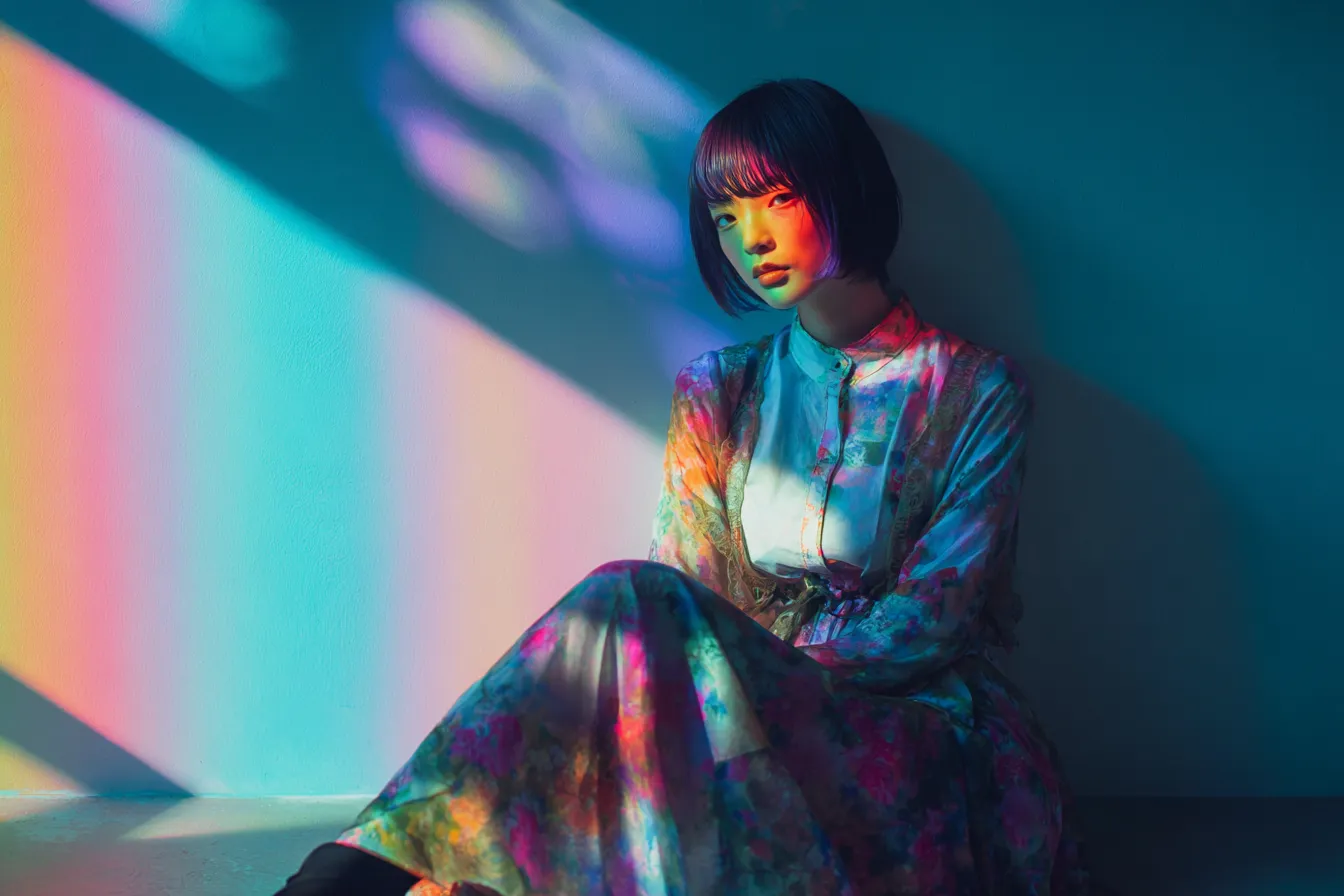 グラデーション背景と座る女性｜壁面に映る虹色の光が空間全体を彩り、グラデーション背景が女性の柔らかさと個性を引き立てるアーティスティックな構図となっている。