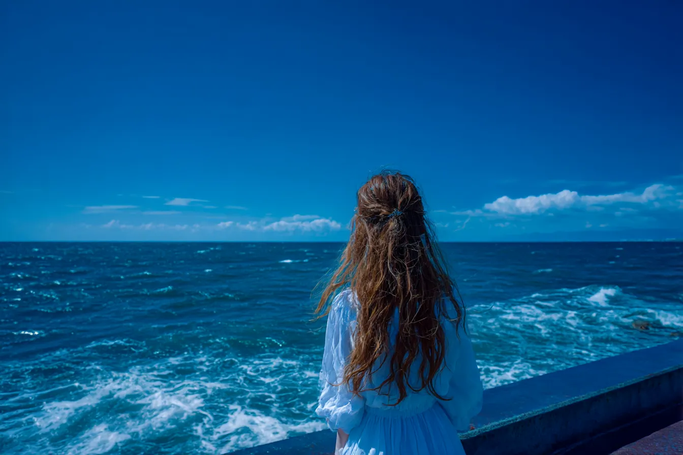 海背景と風になびく茶髪の女性｜強い陽射しと深い青が際立つシーンで、海背景が開放感と旅情を感じさせるドラマティックな印象を与える。
