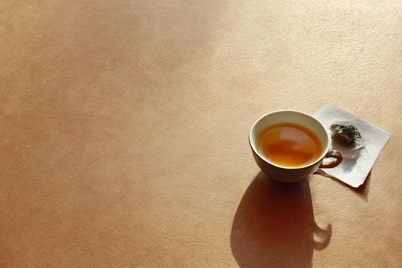 ベージュ背景のティーカップ静物｜光沢感ある茶系の面に、カップと和菓子が対角に置かれた構図。ベージュ背景が温かく穏やかな空間を表現し、落ち着きを感じさせる。