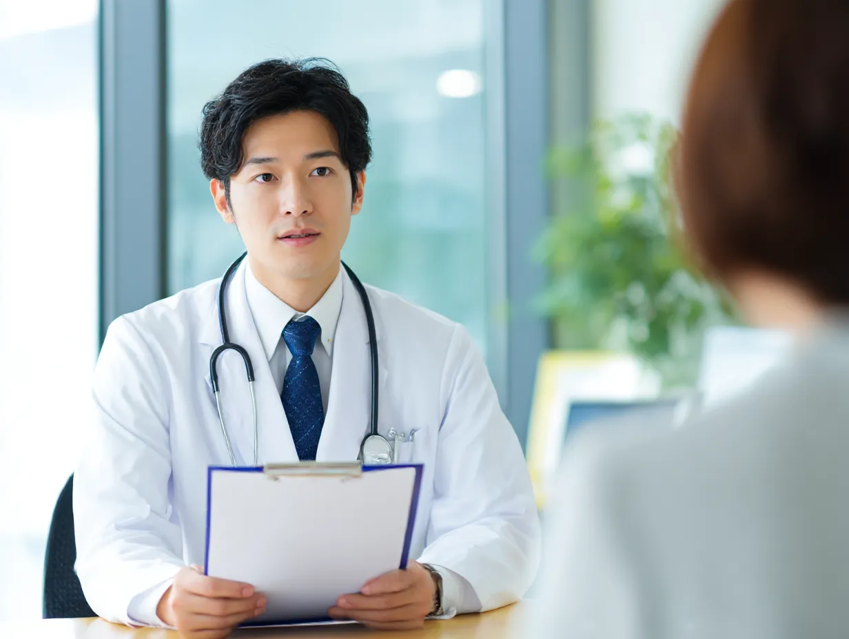 若手男性医師の診察写真｜明るい診察室で若手男性医師がカルテを手に丁寧に話を聞くシーン。清潔感と安心感を与える柔らかい光と構図で、初診や相談の場面に適したイメージ。