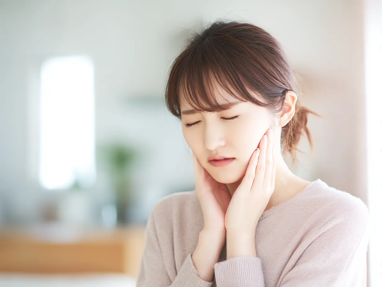 虫歯写真・両頬を押さえる若い女性｜若い女性が室内で両頬を押さえ痛みを感じる様子。柔らかい自然光のなか、表情と仕草が虫歯のつらさをリアルに伝える構図。