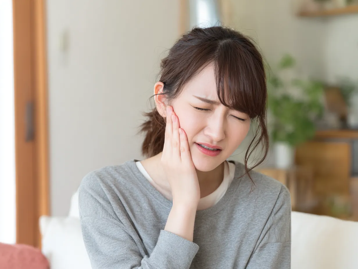 虫歯写真・片頬に痛みを感じる女性｜日常空間で頬に手を当てて痛みに耐える若い女性。暖かみのある室内光が苦しみの中にも生活感を演出するバランスの取れた写真。