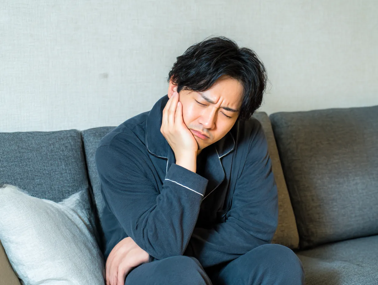 虫歯写真・ソファでうずくまる若い男性｜部屋着姿の男性がソファで顔をしかめている様子。家庭的な背景の中で、虫歯の痛みのリアルさが浮き彫りになる演出。