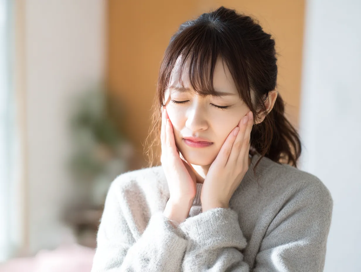 虫歯写真・痛みに苦しむ若い女性｜手を頬にあて目を閉じて痛みに耐える若い女性。室内の優しい光と穏やかな背景が、痛みのコントラストを際立たせる。