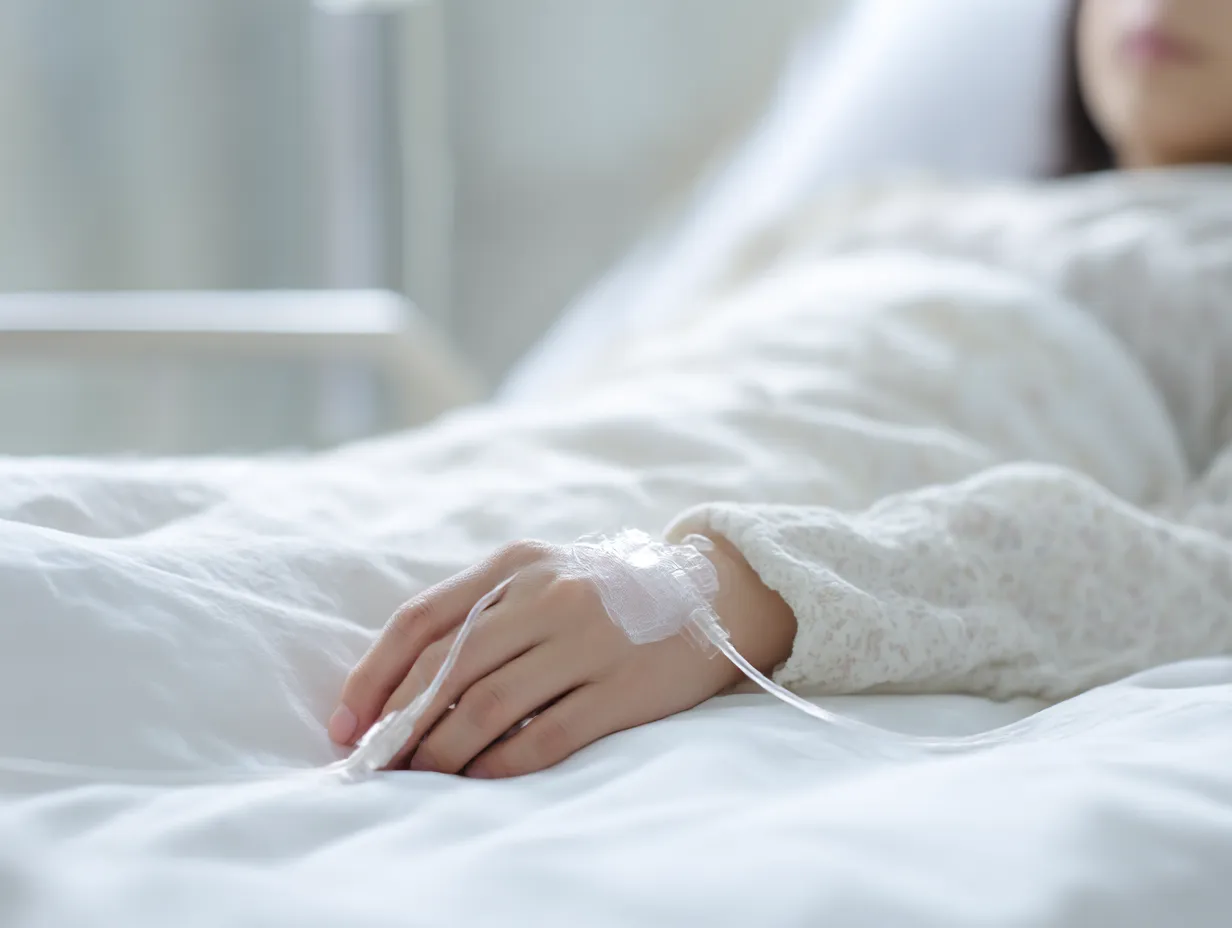 若い女性患者の静かな入院患者写真｜白い寝具と淡い衣類に包まれた若い女性の手元を捉えた入院患者写真。やわらかな光と繊細な描写が、静けさと回復への祈りを伝える。