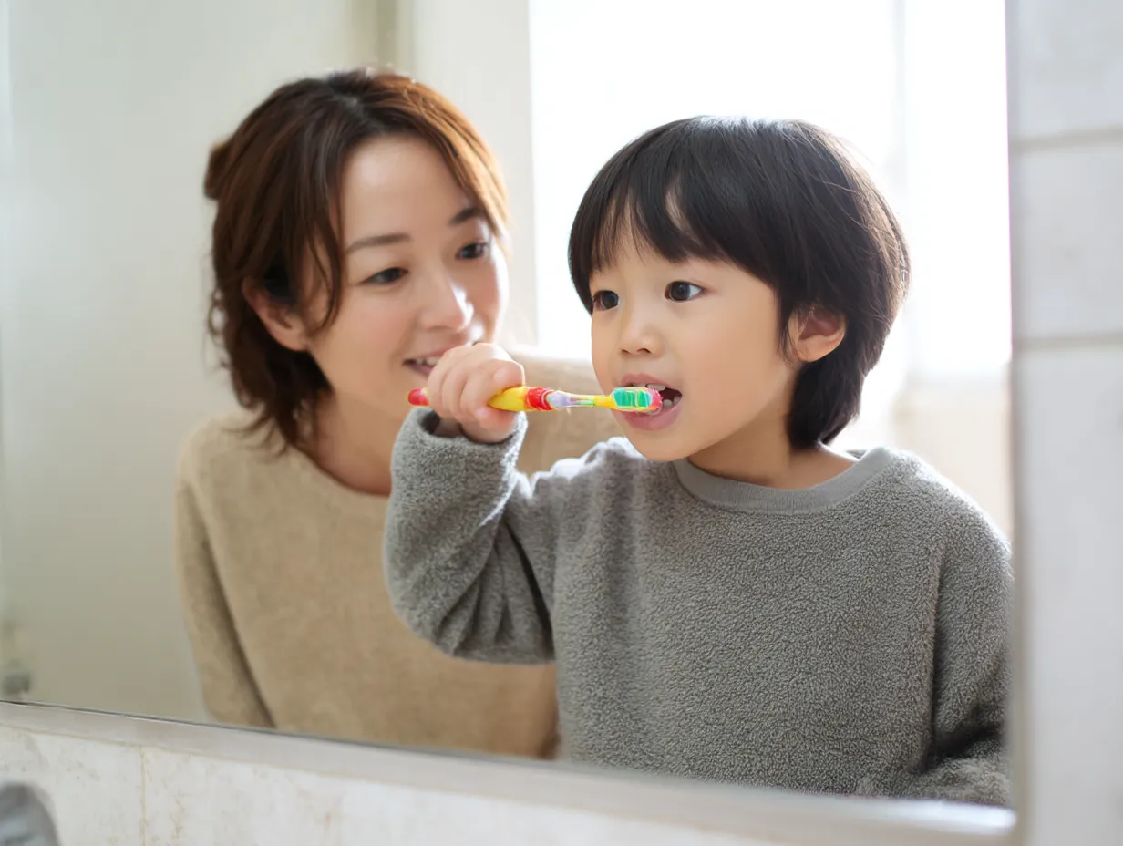 子どもの歯磨き写真と母親の笑顔｜洗面所で母親が見守るなか、真剣に歯磨きをする子どもの姿を捉えた温かな構図。自然光と親子の柔らかい表情が、家庭の健康意識とやさしさを印象づける。