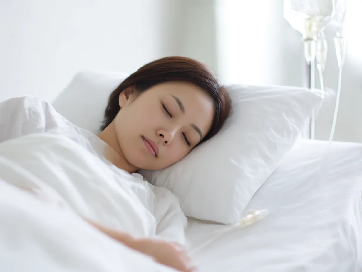 安らかに眠る患者の点滴写真｜白い寝具に包まれ眠る女性と点滴器具が共に写る点滴写真。光と構図が優しさと安心感を醸し、回復への祈りを感じさせる温かな表現。