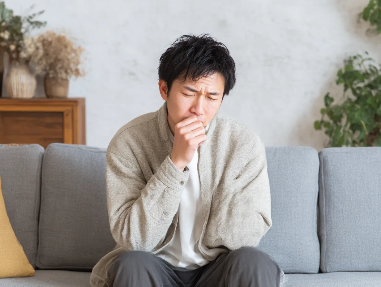 若い男性の喉が痛い写真（室内）｜ソファに腰掛け、咳き込むように口元を手で覆う若い男性。温かみのある室内光と落ち着いた背景が体調不良の辛さをやさしく伝える。