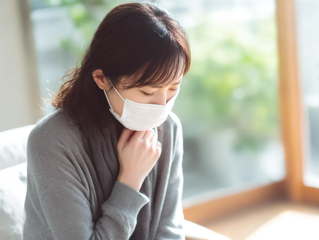 マスク姿の女性の喉が痛い写真｜柔らかな光が差し込む室内で、マスク越しに喉を押さえる女性。健康への不安や感染対策の現代的テーマを静かな画風で表現している。