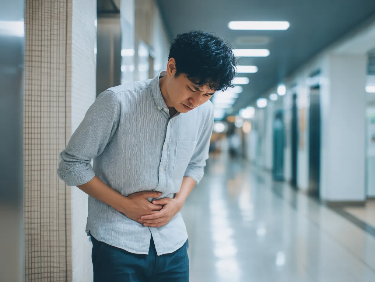 若い男性のお腹が痛い写真｜通路で立ち止まり、両手でお腹を押さえて顔をしかめる若い男性を、青白い蛍光灯の照明が映すシリアスな構図。オフィスビルなどでの急な腹痛をドラマチックに切り取った一枚です。