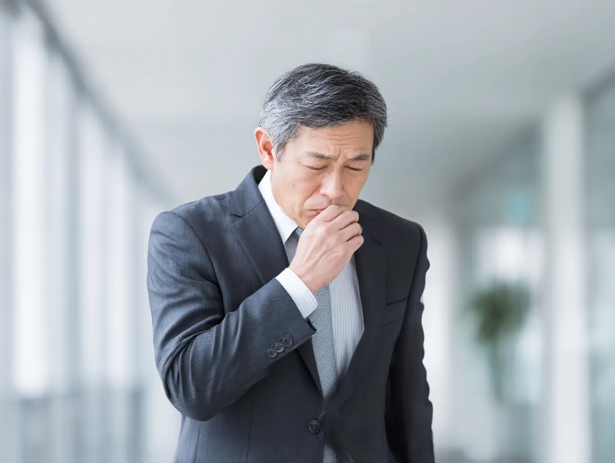 スーツ中年男性の咳写真｜オフィスビルの廊下でスーツ姿の中年男性が咳き込む様子を、白を基調とした明るい背景でクリアに表現。働き盛りの体調変化を静かに浮き彫りにする印象的な構図です。