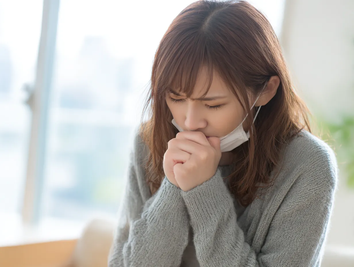 マスク女性のくしゃみ写真｜明るい室内でマスクをずらした若い女性が、くしゃみを抑えるように口元を両手で押さえている姿を、柔らかな逆光で繊細に描写。寒さやアレルギーを思わせる静けさが伝わる画です。